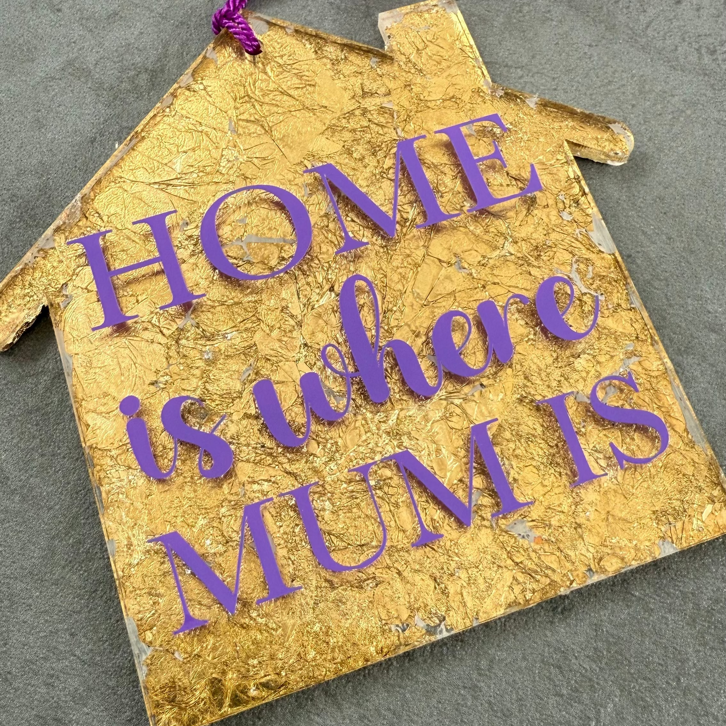 Gold house-shaped ornament with purple text 'HOME is where MUM is' on a gray surface