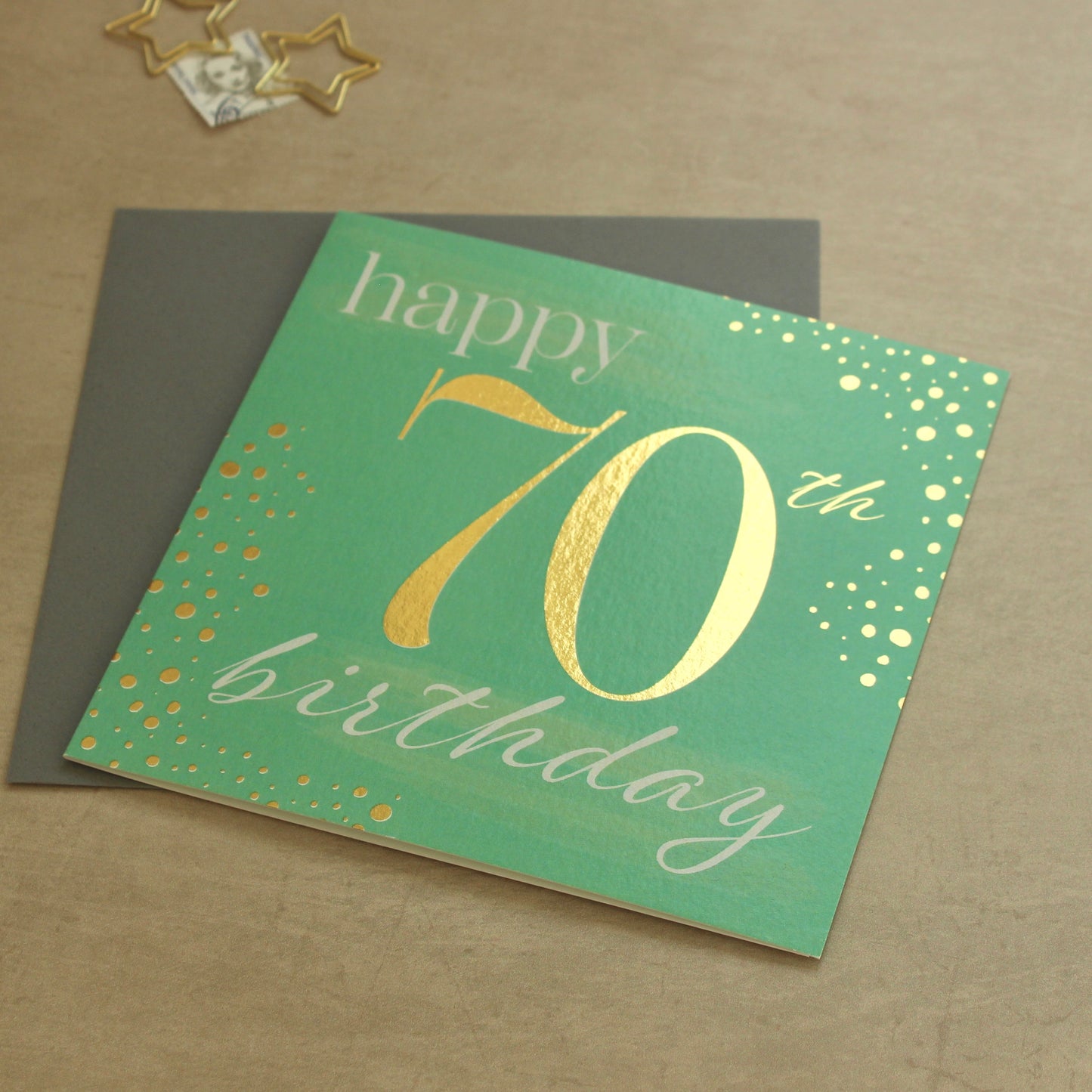 Green birthday card with gold text 'happy 70th birthday' on a brown surface.
