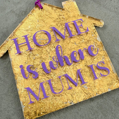 Gold house-shaped ornament with purple text 'HOME is where MUM is' on a gray surface