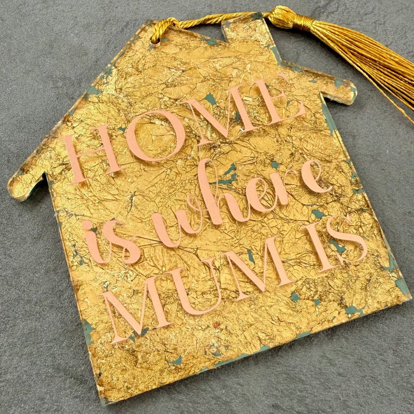 Gold house-shaped ornament with 'HOME is where MUM is' text on a gray surface