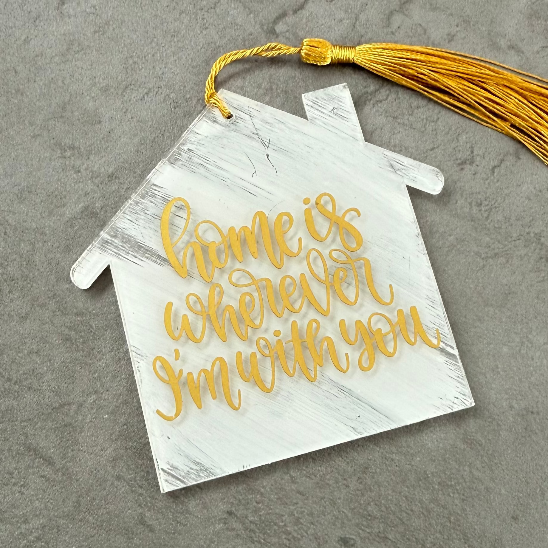 House-shaped decorative tag with 'home is wherever I'm with you' text and a yellow tassel on a gray background