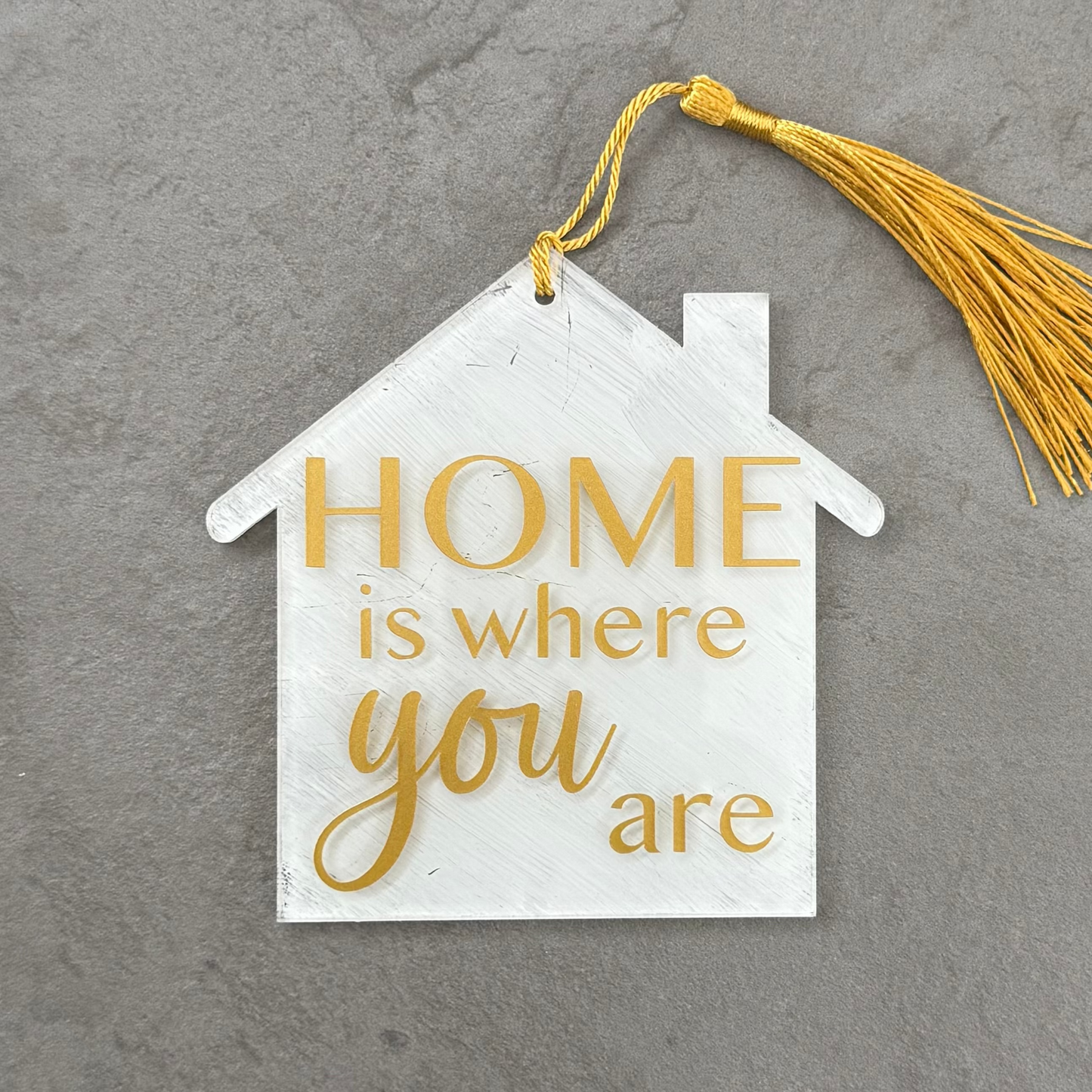 Decorative house-shaped sign with 'HOME is where you are' text and a yellow tassel on a gray background.