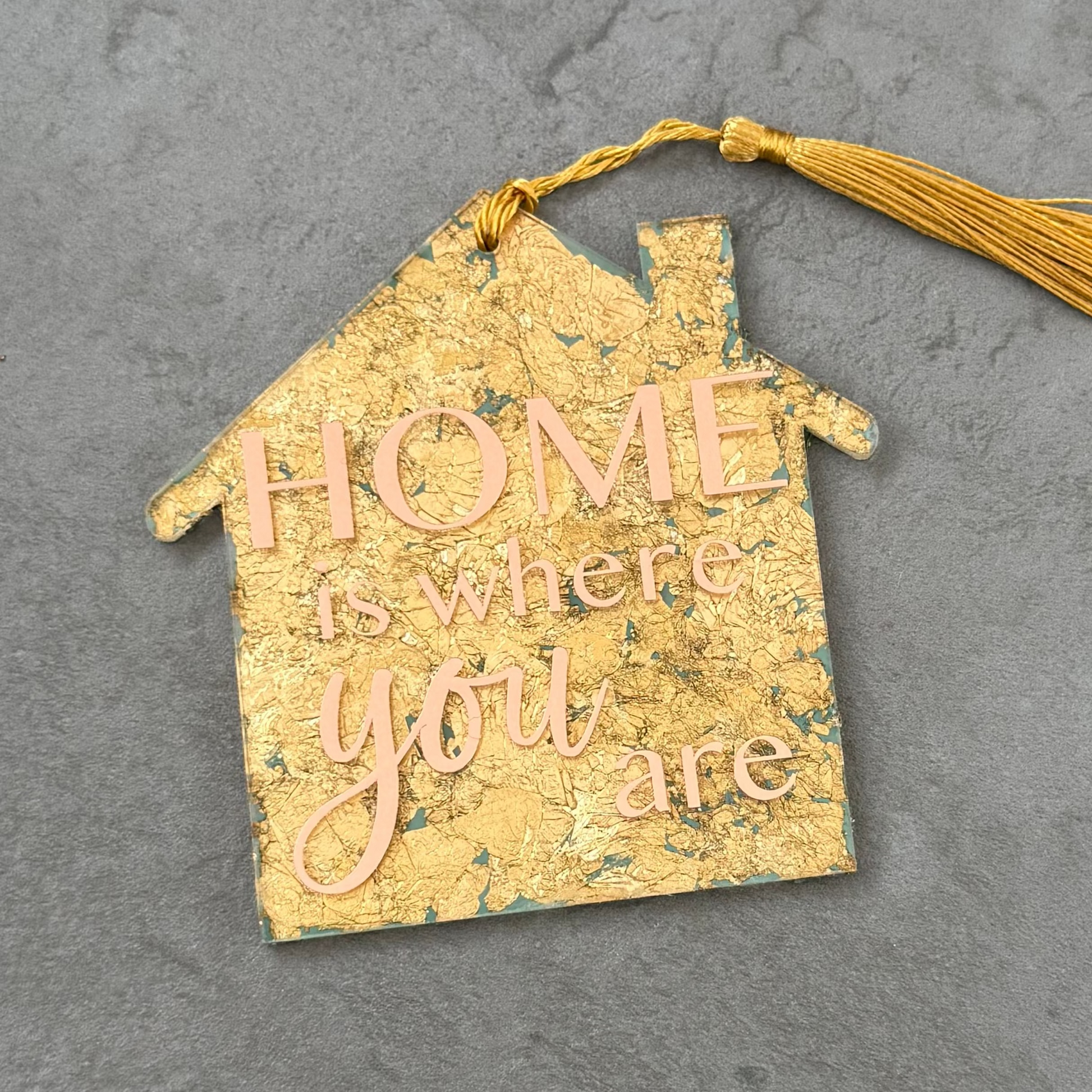 Decorative house-shaped tag with 'Home is where you are' text and a yellow tassel on a gray background.