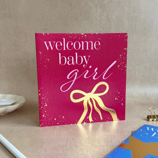 Pink card with 'welcome baby girl' text and gold bow on a beige surface.