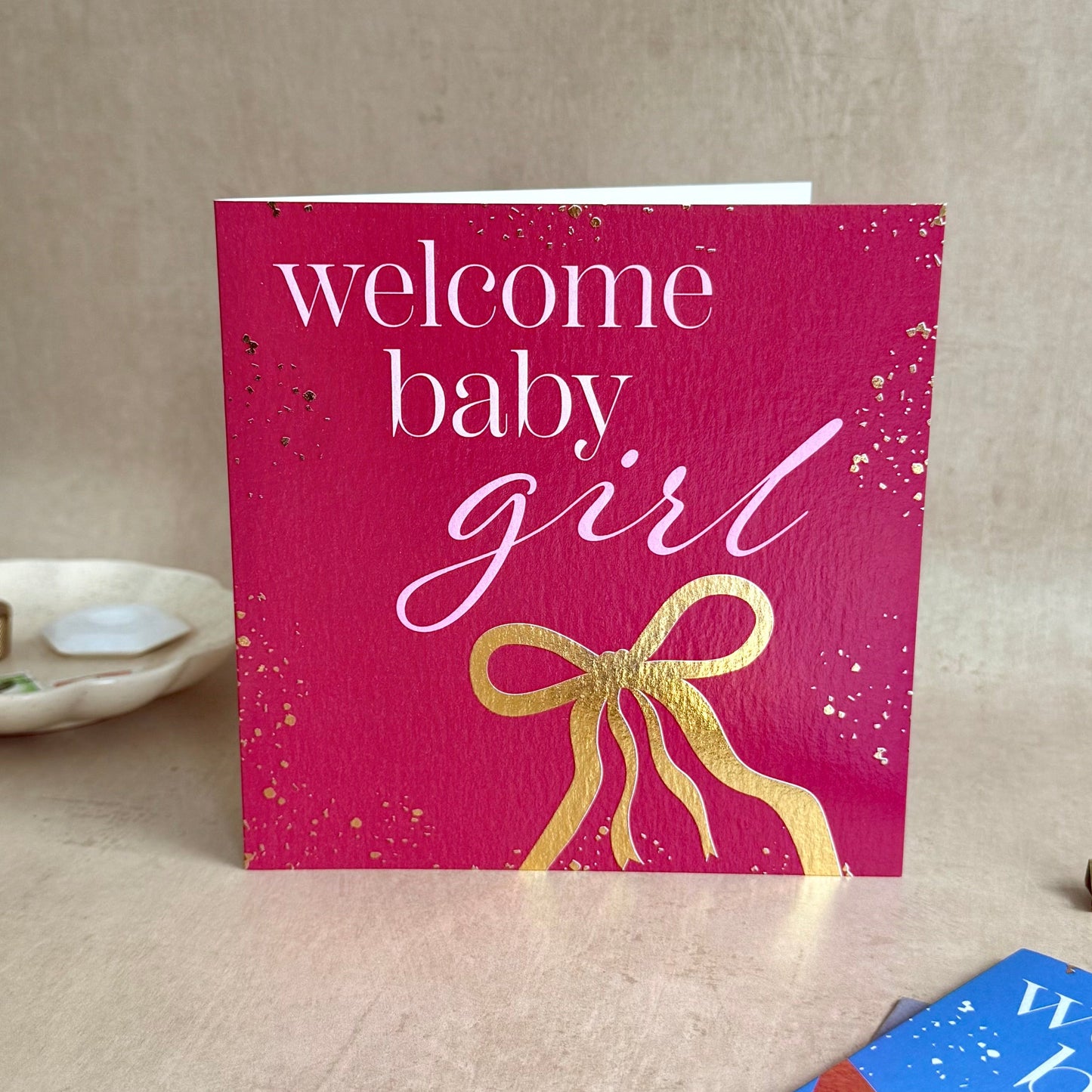 Pink card with 'welcome baby girl' text and gold bow on a beige surface.