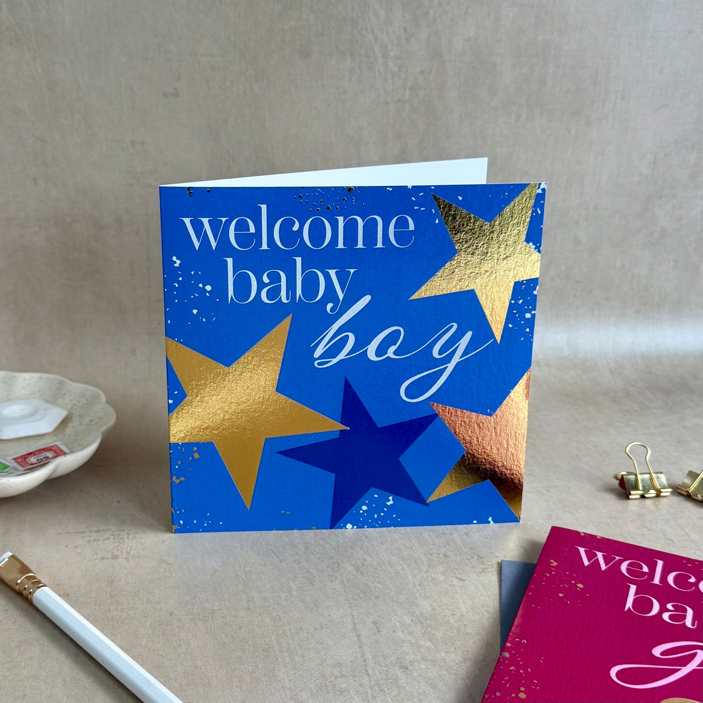 Greeting card with gold and blue stars and 'welcome baby boy' text on a neutral background.