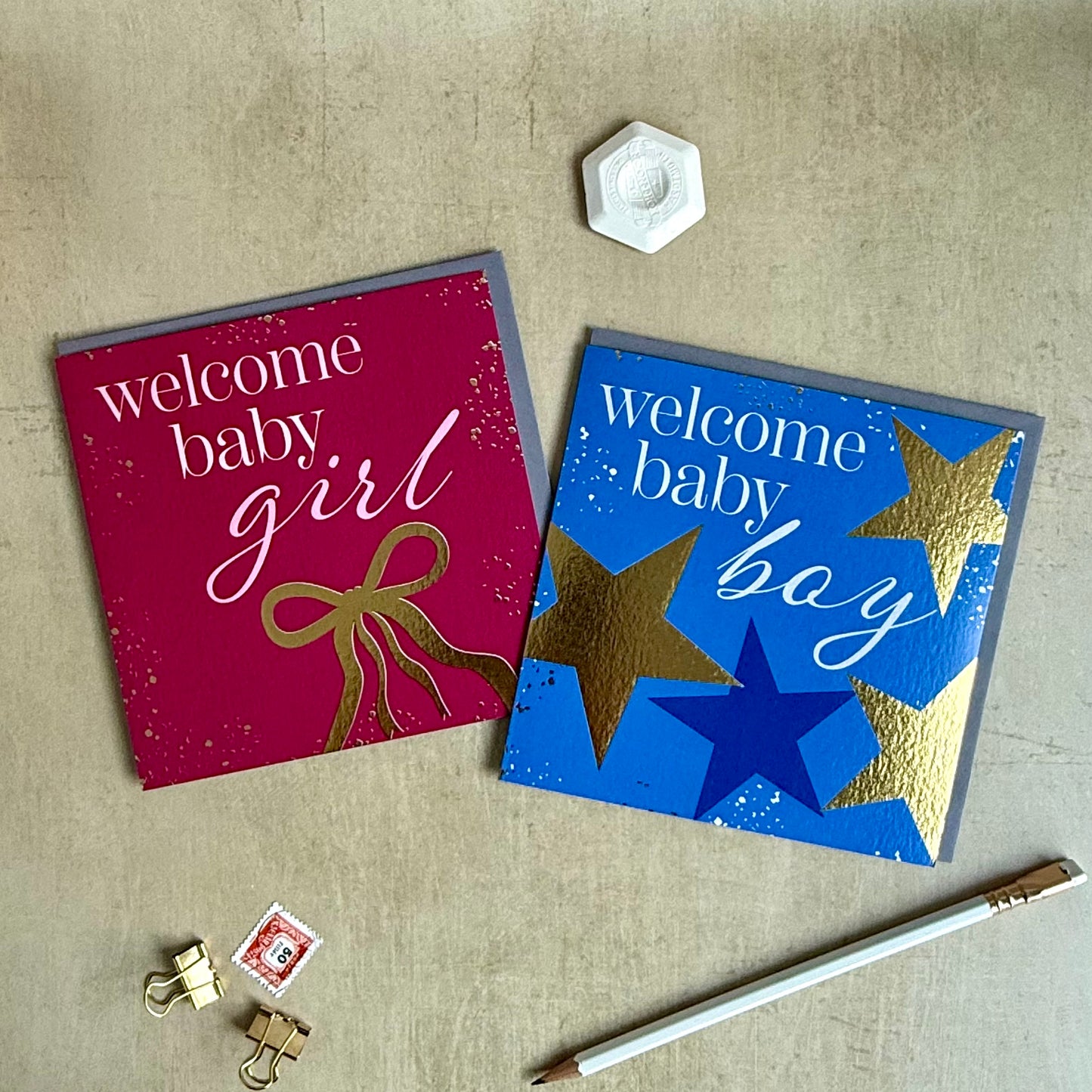 Two greeting cards for a baby shower, one red with 'welcome baby girl' text and bow, the other blue with 'welcome baby boy' text and stars, on a textured surface.