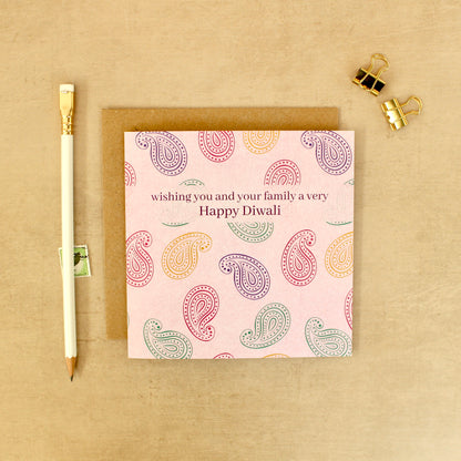 A square greeting card with a pink background and multicolored paisley pattern, placed on top of a brown envelope on a beige surface. The card reads 'wishing you and your family a very Happy Diwali'.