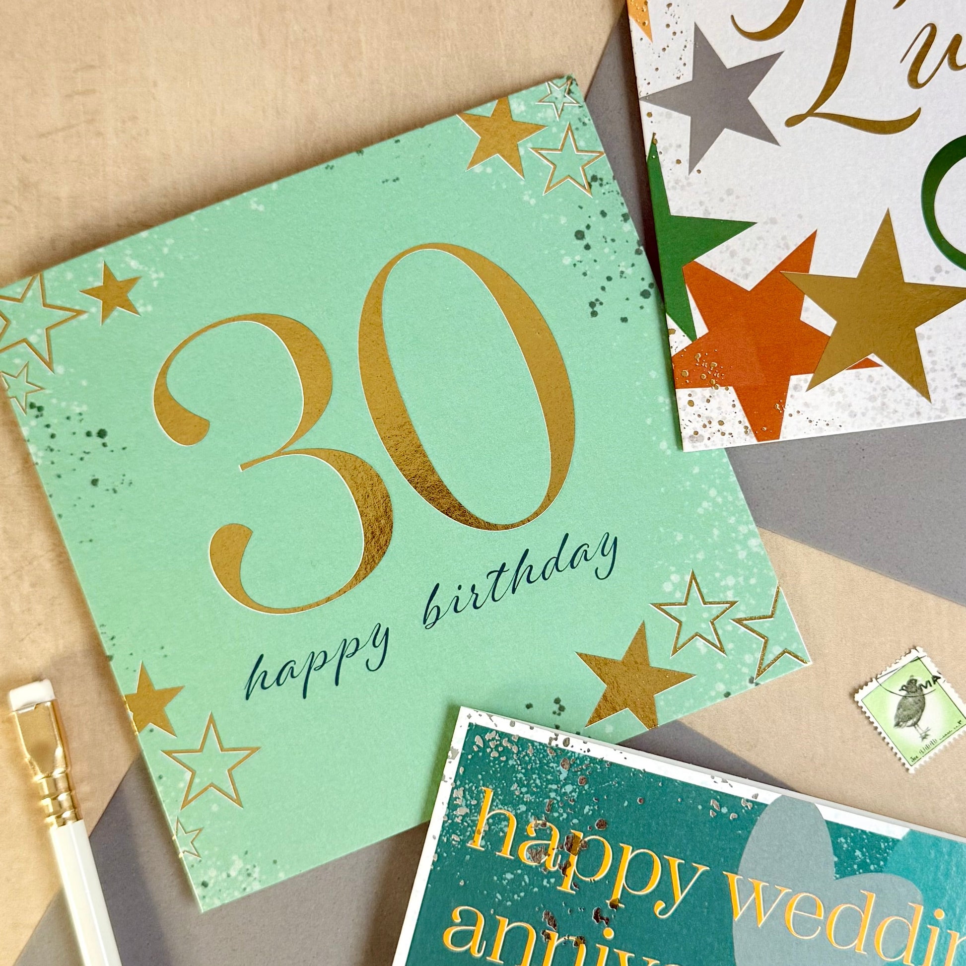 Three greeting cards with '30 happy birthday', 'Good Luck', and 'Happy Wedding Anniversary' text on a beige surface.