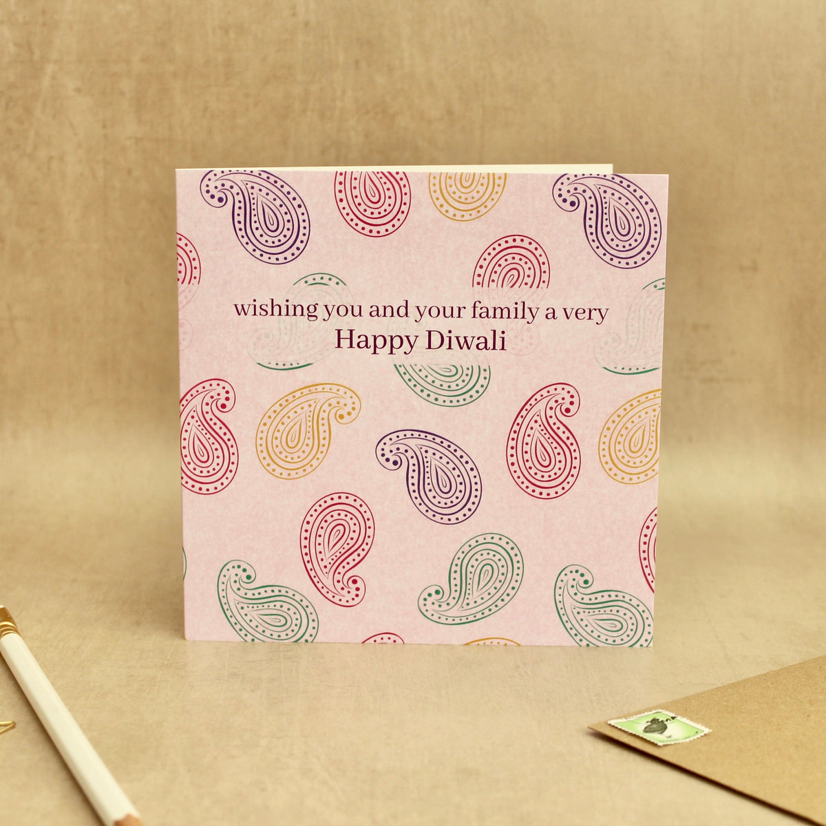A square greeting card with a light pink background and colorful paisley pattern, featuring the text 'wishing you and your family a very Happy Diwali'. The card is displayed on a textured beige surface next to a gold pen, gold star stickers, and a brown envelope.