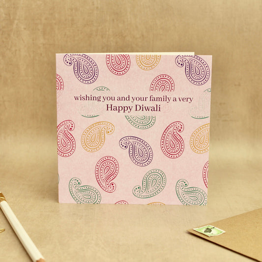 A square greeting card with a light pink background and colorful paisley pattern, featuring the text 'wishing you and your family a very Happy Diwali'. The card is displayed on a textured beige surface next to a gold pen, gold star stickers, and a brown envelope.